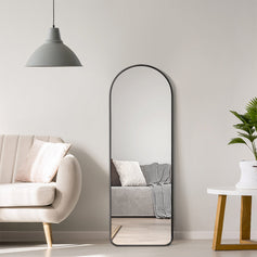 In a modern living room, a black-framed rectangular full-length mirror hangs on a light gray wall, beside a beige sofa and a small side table, showcasing a minimalist and stylish design