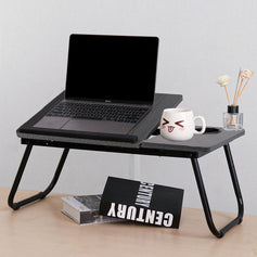 Foldable bed table on a desk with one half lifted to elevate a laptop, next to the other half holding a cup and perfume, two books placed underneath, practical and tidy