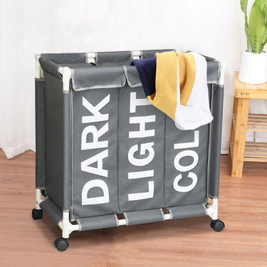 Light grey laundry basket with Velcro lids, three compartments, and clothes hanging from the top, standing upright in the living room